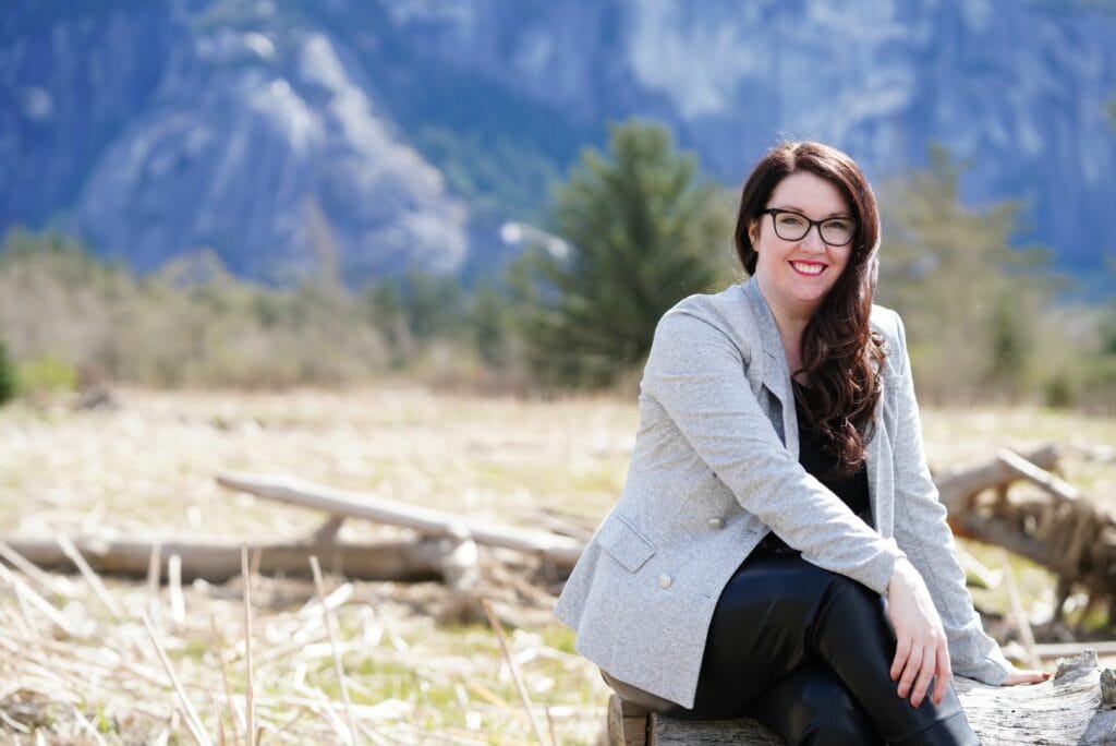 A picture of a caucasian female with dark hair and glasses wearing a grey blazer sitting on a log in the Squamish Estuary, with mountains and tan colored grasses behind her.